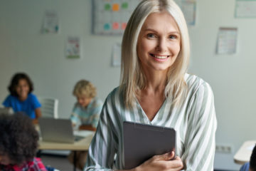 Eine Lehrerin steht mit dem Rücken zu ihrer Klasse und hält ein Tablet in den Händen.