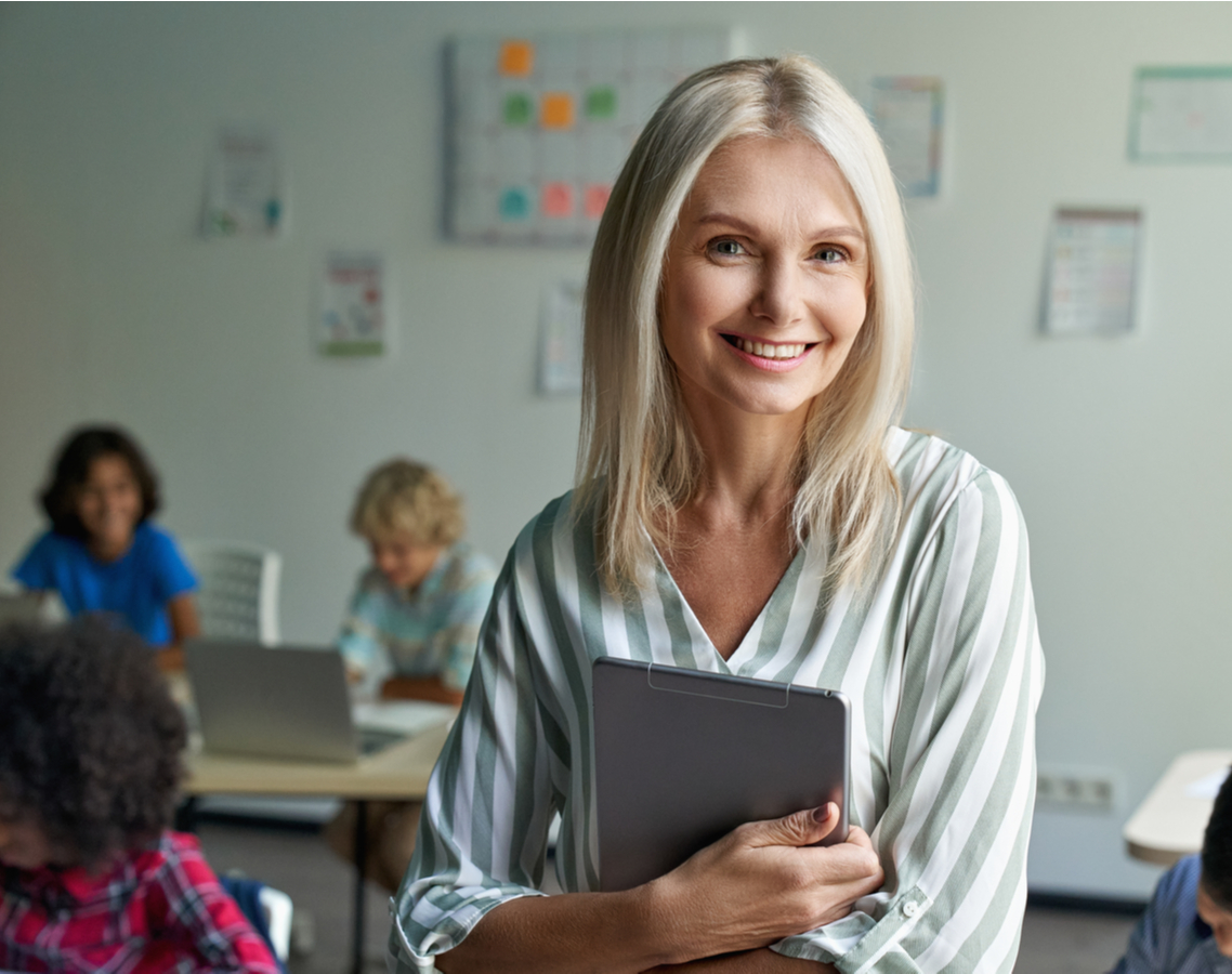 Eine Lehrerin steht mit dem Rücken zu ihrer Klasse und hält ein Tablet in den Händen.