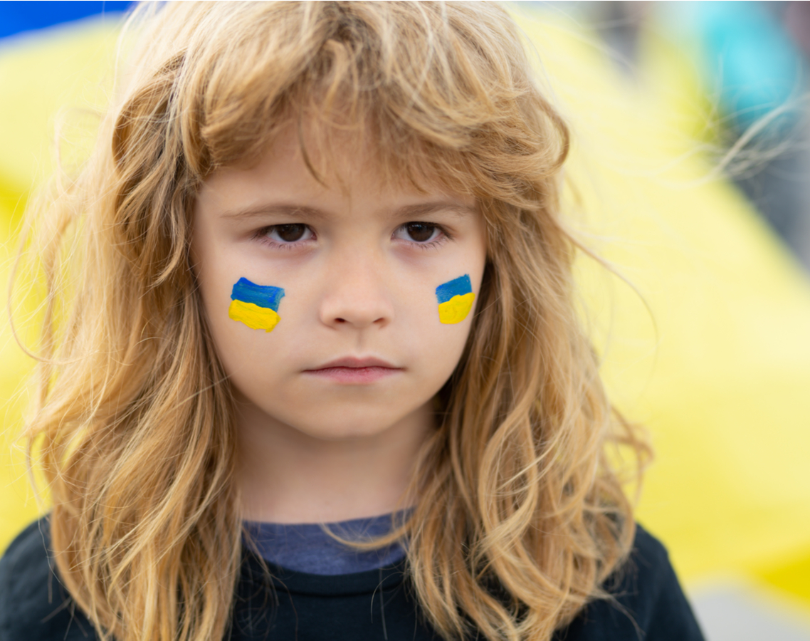 Ein junges Mädchen mit aufgemalten Ukraine-Flaggen auf den Wangen steht traurig guckend vor einer Ukraine-Flagge.