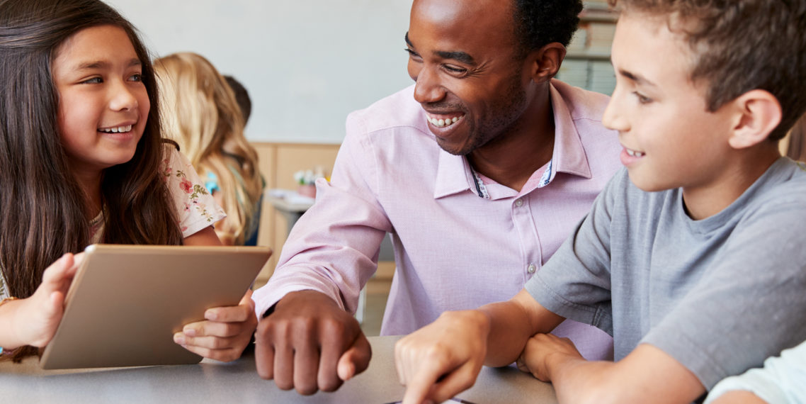 Ein Lehrer im Gespräch mit einer Schülerin und einem Schüler, die beide an einem Tablet arbeiten.