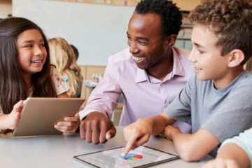 Ein Lehrer im Gespräch mit einer Schülerin und einem Schüler, die beide an einem Tablet arbeiten.