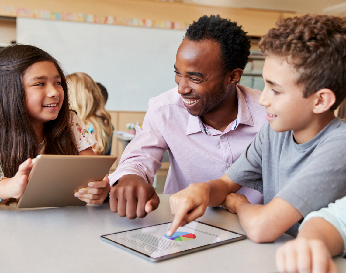 Ein Lehrer im Gespräch mit einer Schülerin und einem Schüler, die beide an einem Tablet arbeiten.