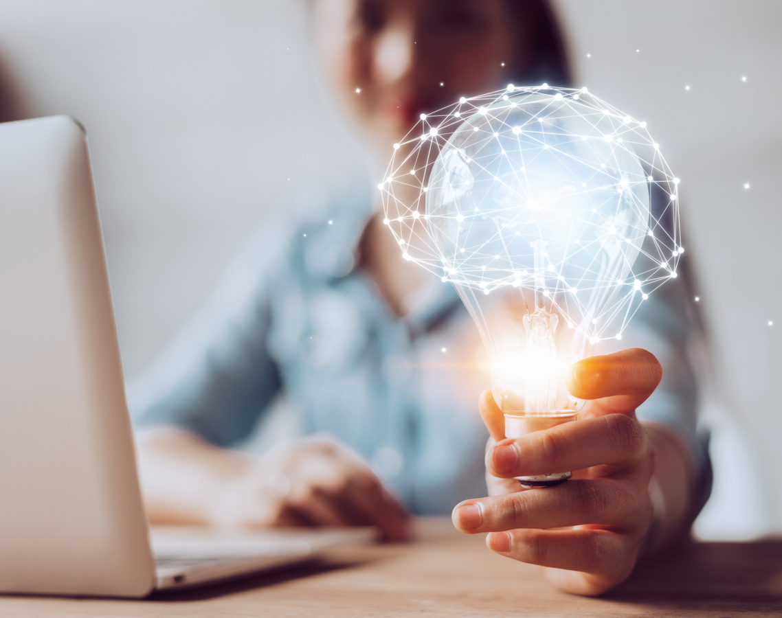 Eine Frau sitzt vor einem Laptop und hält eine leuchtende Glühbirne in die Kamera, über die die Grafik eines Gehirns aus feinen Netzverbindungen gelegt ist.