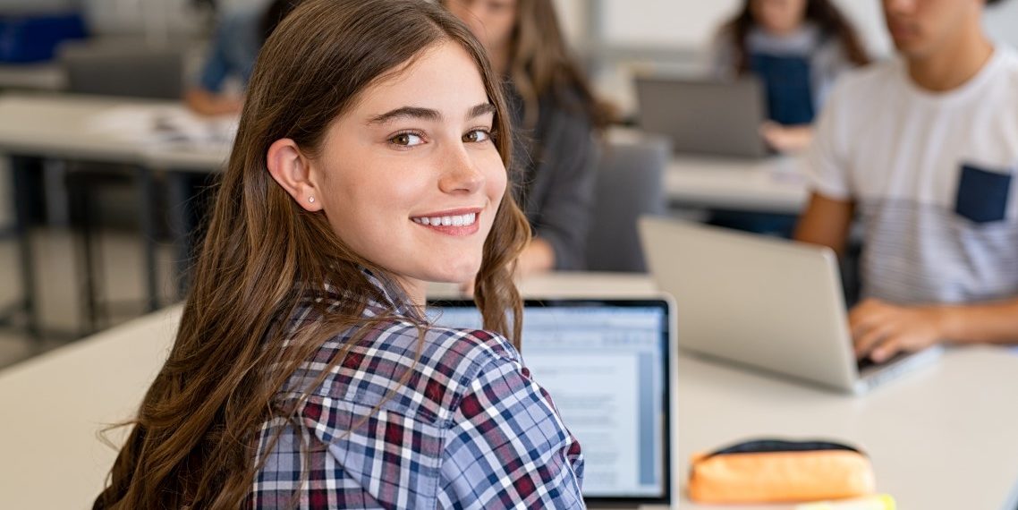 Eine Schülerin arbeitet im Unterricht am Laptop und guckt über ihre Schulter in die Kamera.