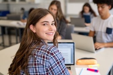 Eine Schülerin arbeitet im Unterricht am Laptop und guckt über ihre Schulter in die Kamera.