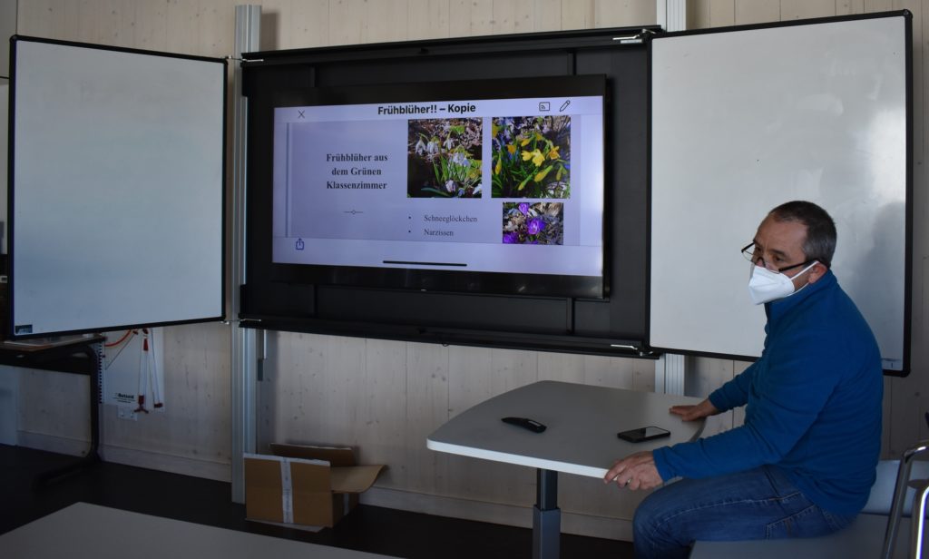 Der stellvertretende Schulleiter der Aspe Realschule, Andreas Wolf, sitzt im Klassenraum vor einer digitalen Tafel, die eine Präsentation über Frühblüher zeigt.