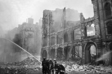 Das schwarz-weiß Foto zeigt eine durch Bomben zerstörte Straße in London, in der fünf Feuerwehrleute versuchen, einen Brand zu löschen.