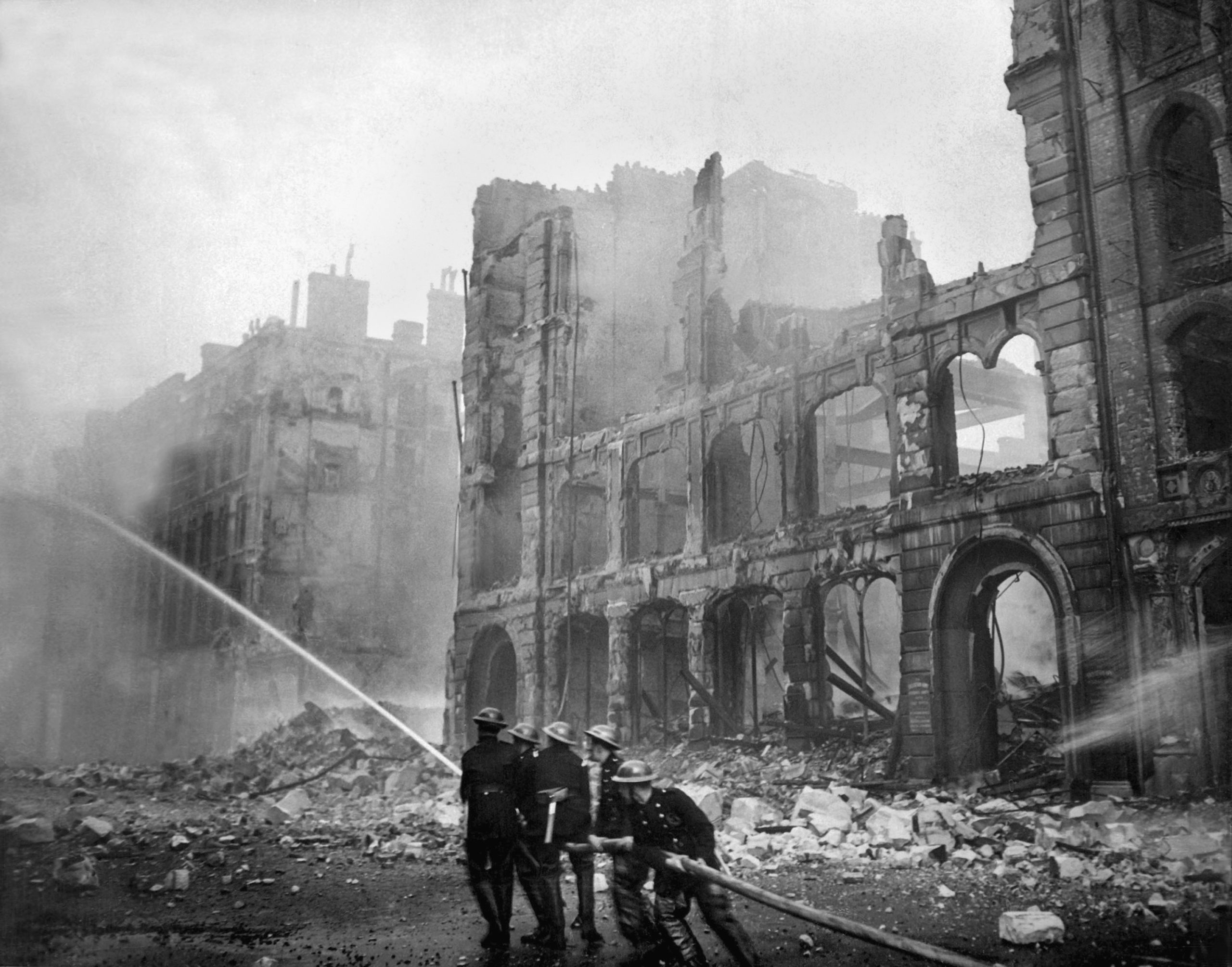 Das schwarz-weiß Foto zeigt eine durch Bomben zerstörte Straße in London, in der fünf Feuerwehrleute versuchen, einen Brand zu löschen.